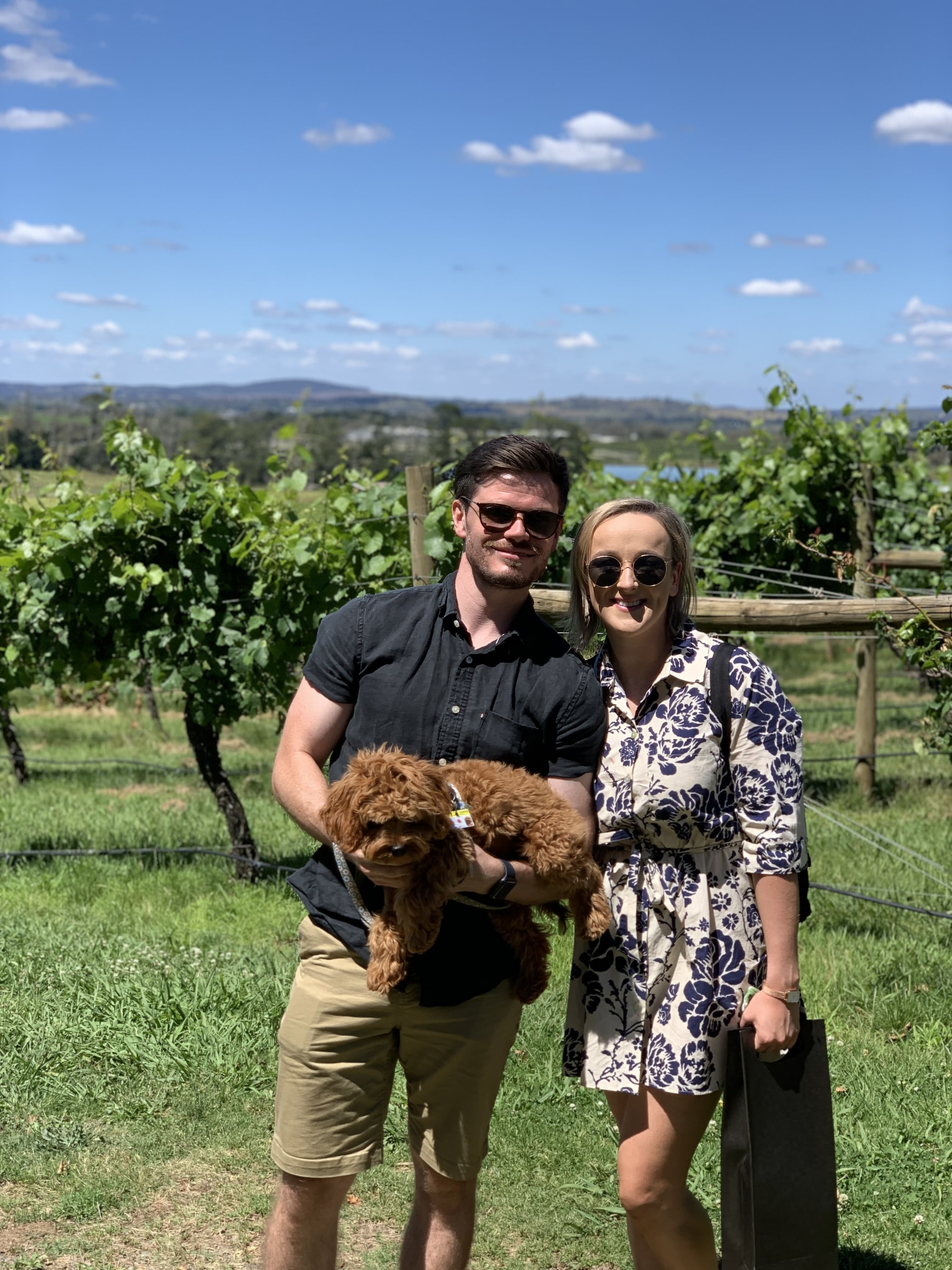 lady and man standing in front of vineyard holding small dog